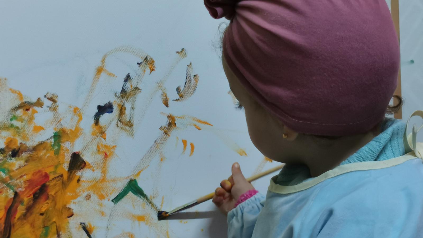 Young child painting on a canvas with colorful strokes, wearing a pink headwrap.